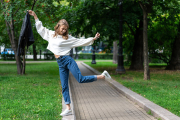 Obraz premium beautiful young European girl walks in city green park. Caucasian girl posing standing on one leg. Lifestyle, pullover, jeans, sneakers. interesting pose full length in fashionable look.