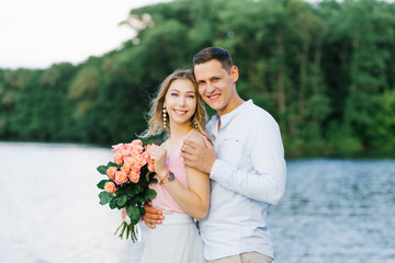 a loving couple a guy and a girl are standing on the Bank of a river or lake. first date. the girl is holding a bouquet of roses, the guy hugs the girl, they smile and