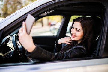 Obraz na płótnie Canvas Happy young woman holding mobile phone and taking photos while driving a car. Smiling girl taking selfie picture with smart phone camera outdoors in car. Holidays and tourism concept