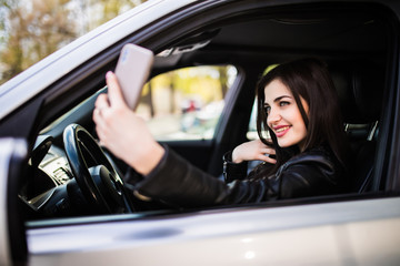 Happy young woman holding mobile phone and taking photos while driving a car. Smiling girl taking selfie picture with smart phone camera outdoors in car. Holidays and tourism concept