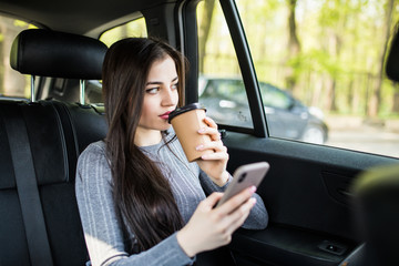 Young woman sitting in the back seat of the car with phone in hand and drinking coffee
