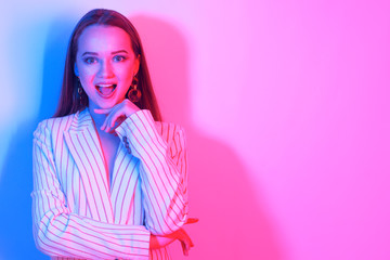 Fashion portrait of young elegant girl in a jacket. Colored neon background, studio shot. Beautiful brunette woman. Hipster girl with long hair surprised, smiles and looks away at copy space