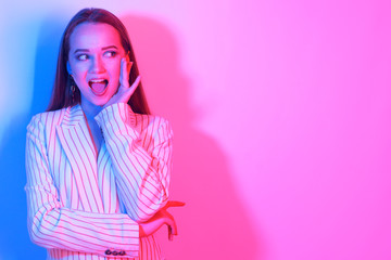 Fashion portrait of young elegant girl in a jacket. Colored neon background, studio shot. Beautiful brunette woman. Hipster girl with long hair surprised, smiles and looks away at copy space