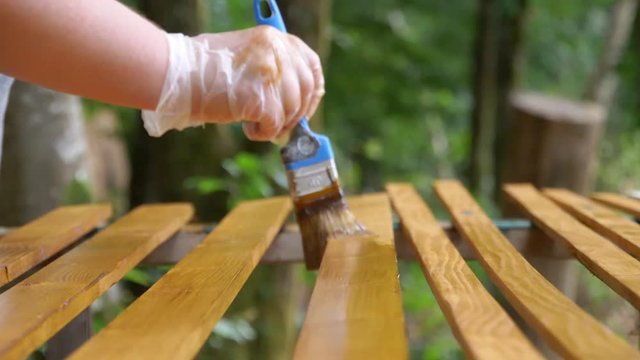 Slow motion hand painting a wood piece in light brown color. The brush goes in and out of focus. Forest in background