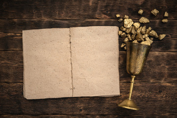 Blank page open book with a copy space and goblet full of gold on a brown wooden table background.