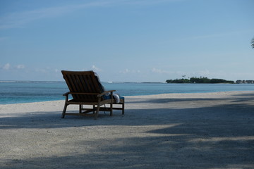 Outdoor deck chair in sunshine shadows on white beach in Maldive island resort.  Sunny blue sky and ocean horizon. relaxing sunbathe concept