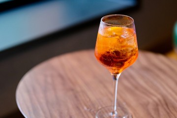 close up a wine glass of orange color cocktail with ice cubes inside on patterned wooden table. Blur background