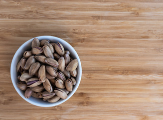 Pistachio nuts poured into the top of a white bowl on a light brown background, top view, on the right is an empty space