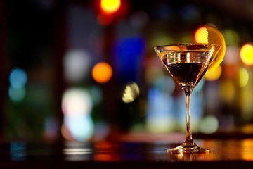 Close up a glass of martini cocktail with orange slice on table in the bar. Defocused colorful lights background