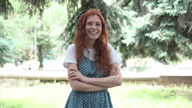 Ginger girl with big headphones wagging her finger laughs and act the maggot. Red-haired woman monkey and play tricks in the park close-up. Freckled teenager outdoor. 