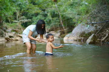 Fototapeta premium Asian mother in blue shirt with DIY chest pocket playing water with her son about 1 year and 9 months at waterfall
