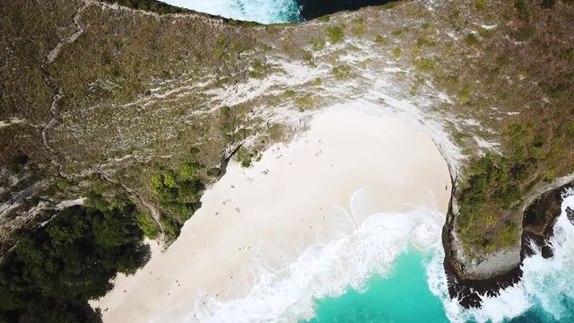 Drone shot flying directly above KelingKing beach on the island of Nusa Penida, Indonesia. The incredible blue water can be seen in waves crashing upon the shore.