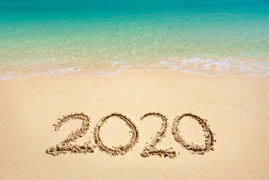 Happy New Year 2020, lettering on the beach with wave and clear blue sea. Numbers 2020 year on the sea shore, message handwritten in the golden sand on beautiful beach background. New Years concept.