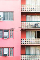 The side of a pink building  with pink walls and ornate white railings and curtains within the sliding door of the apartments, which  lead out to a balcony, hotel, apartment building.