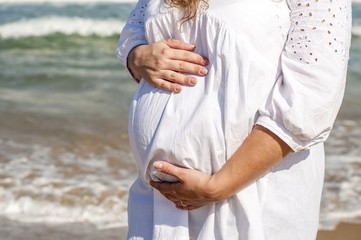 Pregnant young woman in a white dress standing at the seaside and holding her belly with hands.