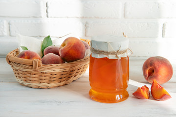 Jam and peaches in a wicker basket on a white wooden background. Morning sun on a white brick wall.