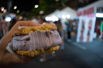 A young woman holds a lavender flavour and purple colored, snickerdoodle cookie ice cream sandwich with a bite taken out of it at a street fair in waikiki at night with bokeh lights in the background.