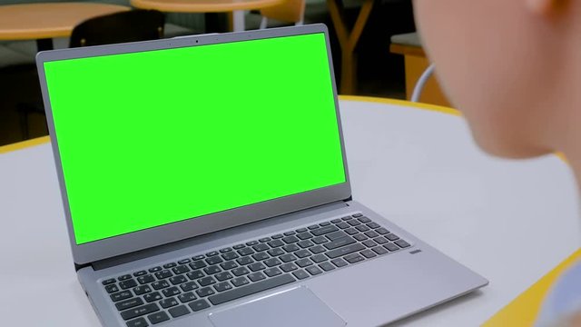 Woman sitting at table and looking at grey laptop computer device with blank green screen in cafe. Technology, chroma key, template, mockup and entertainment concept