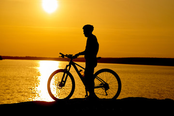 Fototapeta premium Silhouette of a male cyclist with helmet at sunset near the river. Healthy active lifestyle.