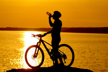 Obraz premium Silhouette of cyclist. Sporty guy cycling in helmet drinks water on the natural background of setting the sun in the evening.