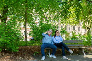 Couple sitting on bench of a city park. Each one is speaking on the phone