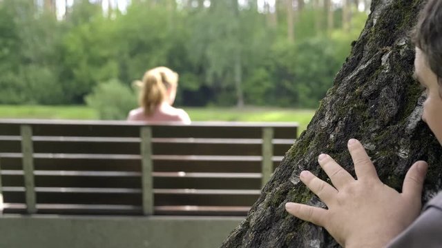 Maniac is watching girl sitting on park bench. Close up portrait of man peeping and looking from behind tree.