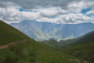 Obraz premium Road leads to the large scenery of rocky mountains, green hills and cloudy sky. Trinity church. Kazbegi Georgia