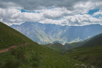 Road leads to the large scenery of rocky mountains, green hills and cloudy sky. Trinity church. Kazbegi Georgia