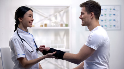 Professional physician fixing titan wrist brace, smiling to male patient, clinic