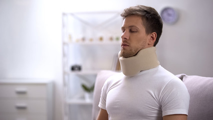 Upset man in foam cervical collar sitting on sofa, accident victim, neck trauma