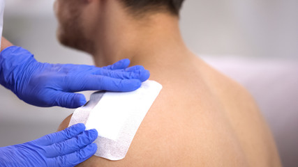 Doctor hands applying adhesive bandage on injured shoulder of male patient - obrazy, fototapety, plakaty