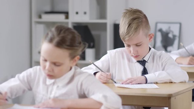 Tracking rack focus shot of multiethnic schoolchildren, wearing uniform white shirts, sitting at their desks in classroom and filling out test answer sheets, looking down at papers and thinking