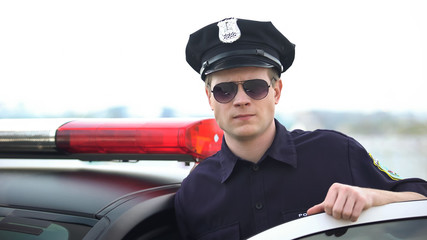 Confident police officer in uniform and sunglasses standing near patrol car