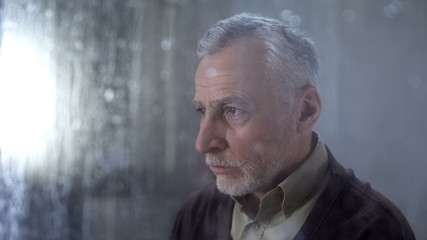 Fototapeta premium Senior man feeling sad, rain drops on window glass, depression concept, lonely