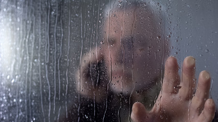 Worried elderly man looking through rainy window, talking on phone, waiting
