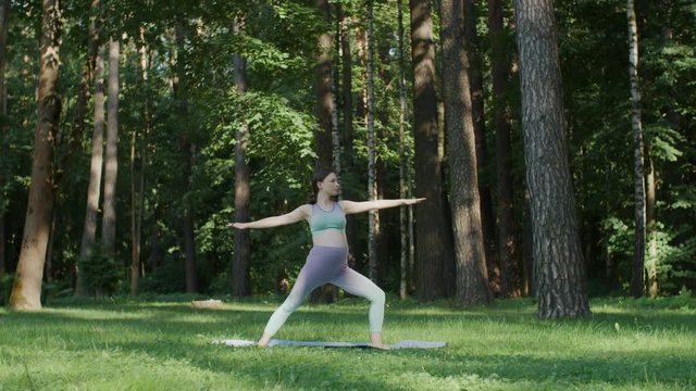 Pregnant woman doing yoga standing in warrior position outdoors on sunny day. Mother to be practice stretching yoga on mat in green park with tall trees on background. Slide camera movement
