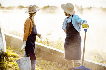 Two well-dressed farmers with working tools standing on a farmland with automatic watering on a farm during the sunset