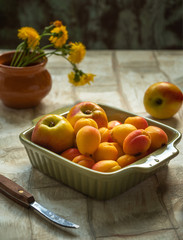 Ripe nectarines and apricots in a ceramic square plate of olive color and a small bouquet of mother-and-stepmother on a light table, a knife for cutting fruit with a wooden handle