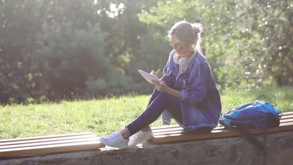 inspired blond girl in denim dressing writes in notebook and thinks sitting on wooden bench in green park slow motion