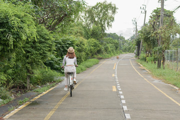 Fototapeta premium Pretty girl with straw hat is happy riding with bike down wide beautiful park alley with trees around on sunny summer day.