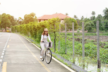 Pretty girl with straw hat is happy riding with bike down wide beautiful park alley with trees around.