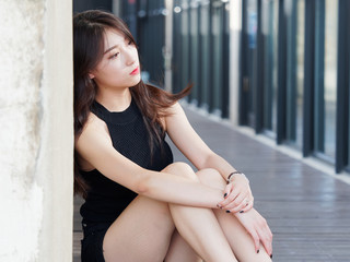 Graceful young woman with long black hair sitting on ground and leaning on the wall in sunny day. Outdoor portrait of cool Chinese girl in trendy summer sexy black dress and sandal, summer concept.