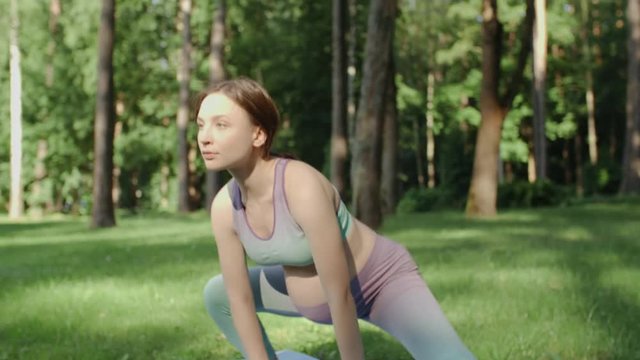 Pregnant woman doing yoga in nature outdoors on sunny day. Young future mother practice stretching yoga in in low lunge position on mat in green park with tall trees on background. Handheld camera