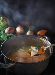 Meat soup with fresh vegetables cooked on the stove. Hot soup in meat broth in a saucepan with handles. Steam over a pot of soup. Raw vegetables are on the table. Сooking dinner