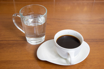 A cup of black coffee on a saucer and a glass of water on a wooden table