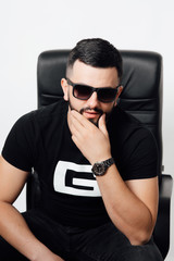 Obraz premium Charming and stylish. Perfect style. Handsome young man keeping hand on chin and looking at camera while sitting against white background. Fashion man posing.
