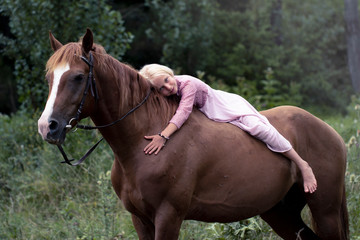 blonde girl with horse in the forest. beautiful Caucasian girl with long blonde hair in a pink dress lies on a brown horse and hugs her. barefoot child on horseback in the forest Bareback.