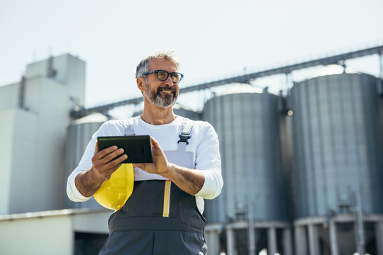 Engineer Using Tablet In Front Of Blurred Grain Silos