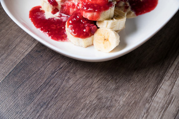 Closeup Vanilla ice cream poured with strawberry or raspberry jam with sliced banana slices on a white plate on a dark wooden table. You can do this at home on vacation