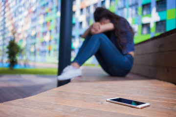 Smartphone lying in the foreground, on blurred background, girl, hiding face in her knees, sitting on a wooden bench. - obrazy, fototapety, plakaty